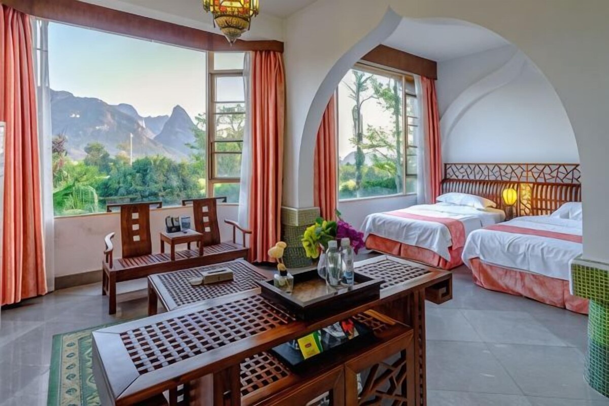 A room with two beds, a dining table, a chair, and a view of mountains.