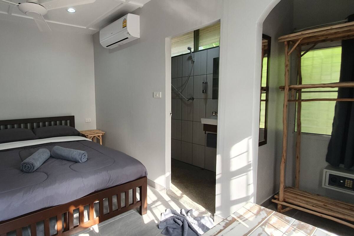 A bedroom with a bed, a wooden headboard, a small wooden stool, and a shower area visible through an open door.