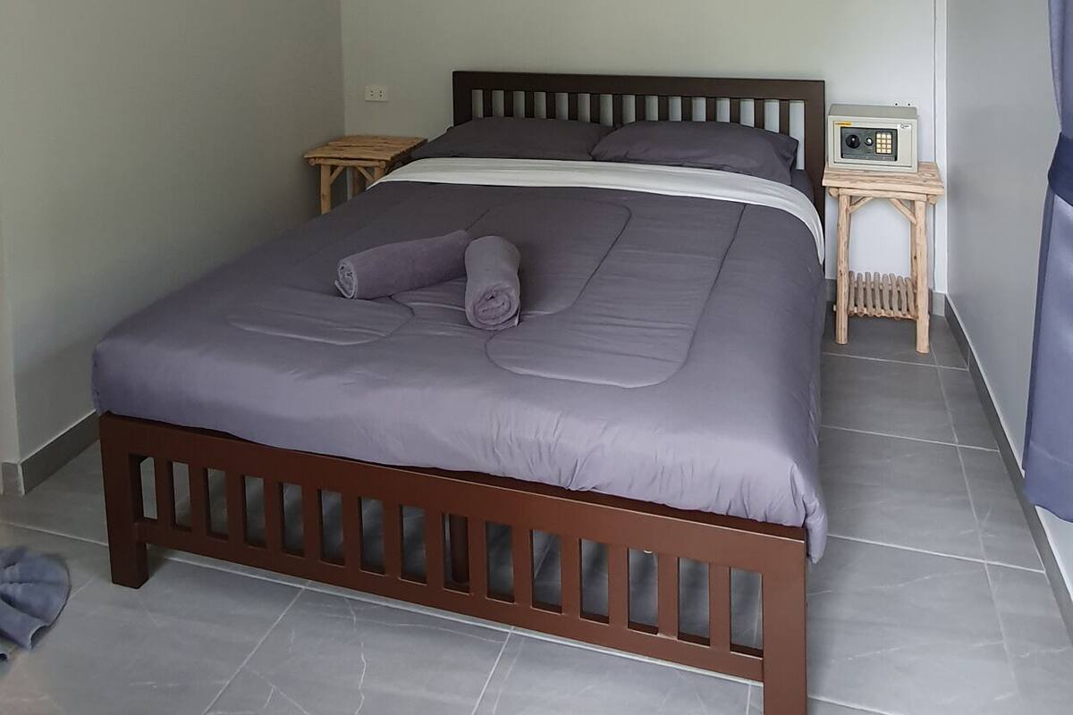 A bedroom with a bed, a ceiling fan, an air conditioning unit, a bedside table with a radio, and a small wooden stool.