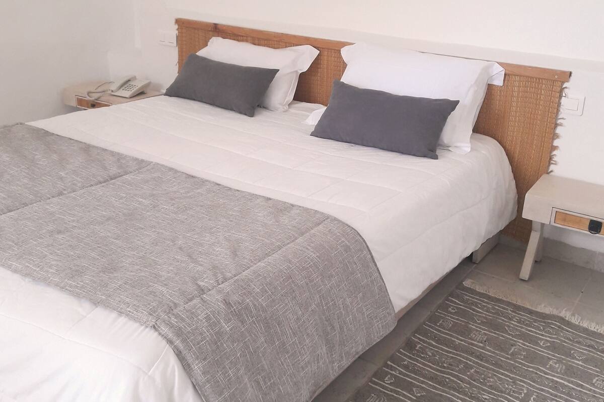 A neatly made bed with a grey blanket and two pillows, a bedside table with a phone, and a painting on the wall.