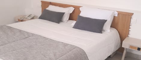 Una cama bien hecha con una manta gris y dos almohadas, una mesita de noche con un teléfono y un cuadro en la pared.