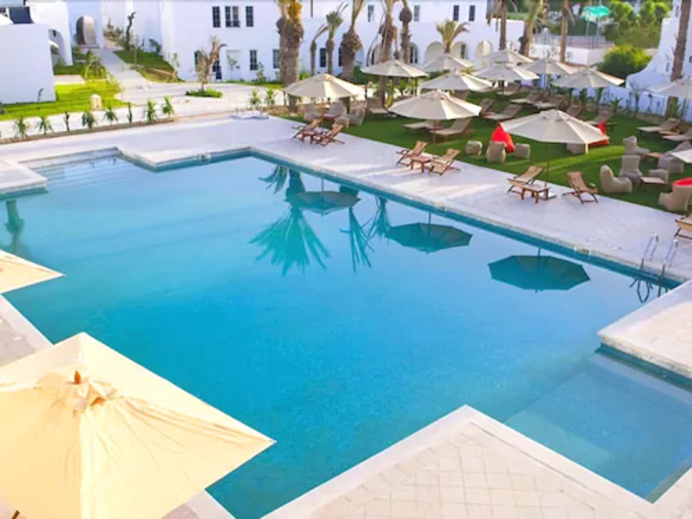 A swimming pool with lounge chairs and umbrellas, surrounded by white buildings and palm trees.