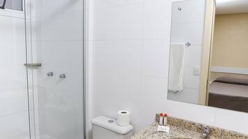 A modern bathroom with a glass-enclosed shower, a white toilet, a granite countertop sink, and a mirror reflecting a bed.