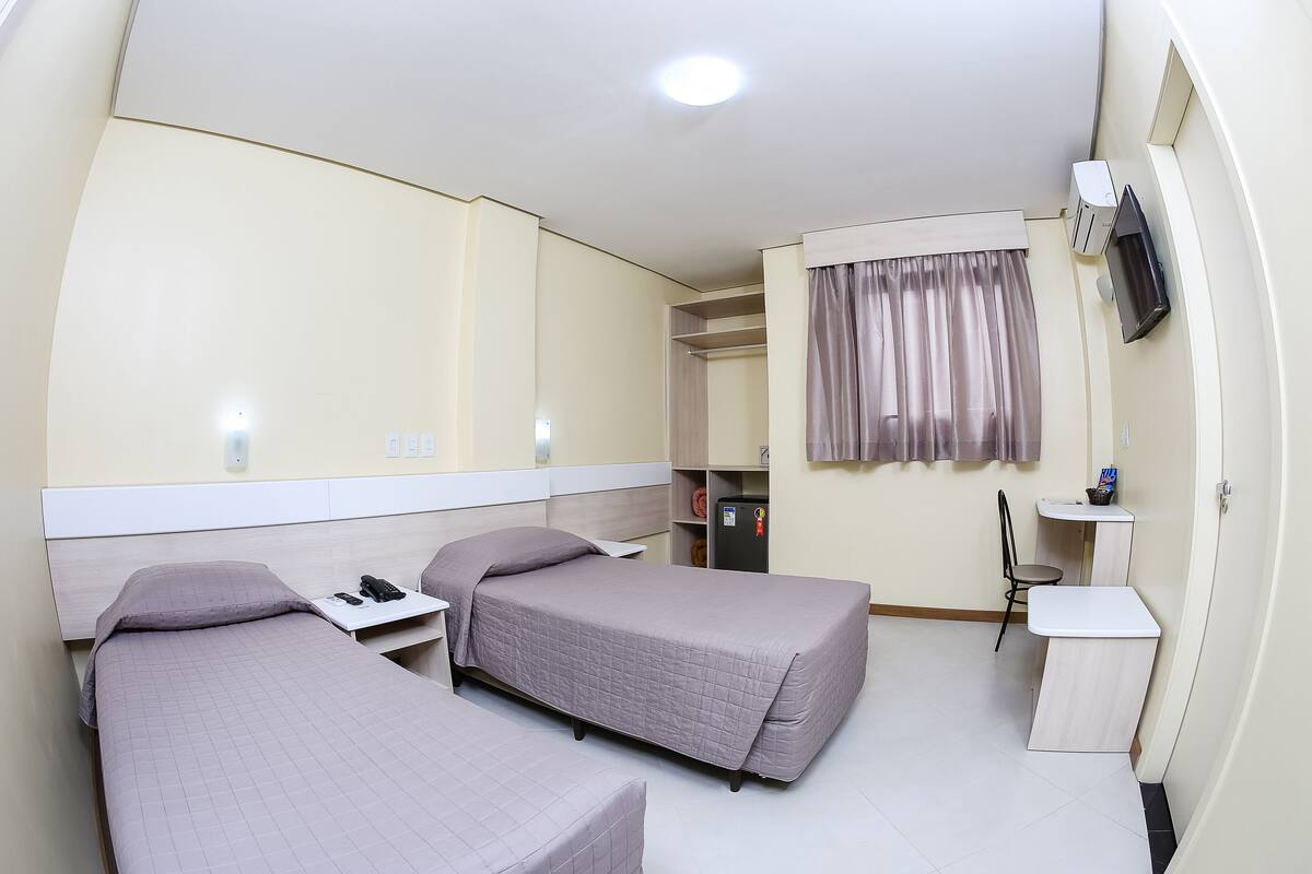 Ein Hotelzimmer mit zwei Einzelbetten, einem Schreibtisch mit Stuhl, einem kleinen Kühlschrank, einem an der Wand montierten Fernseher und einem Fenster mit Vorhängen.