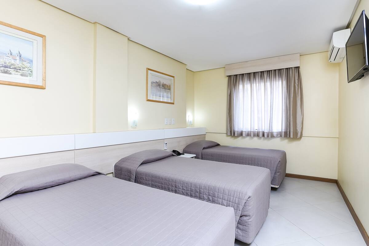 Ein Hotelzimmer mit drei Betten, einem Flachbildfernseher, einer Klimaanlage und einem mit Vorhängen versehenen Fenster.