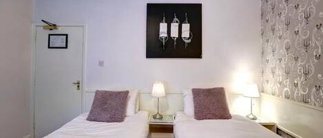 A hotel room with two single beds, a small desk, a lamp, and a framed picture on the wall.