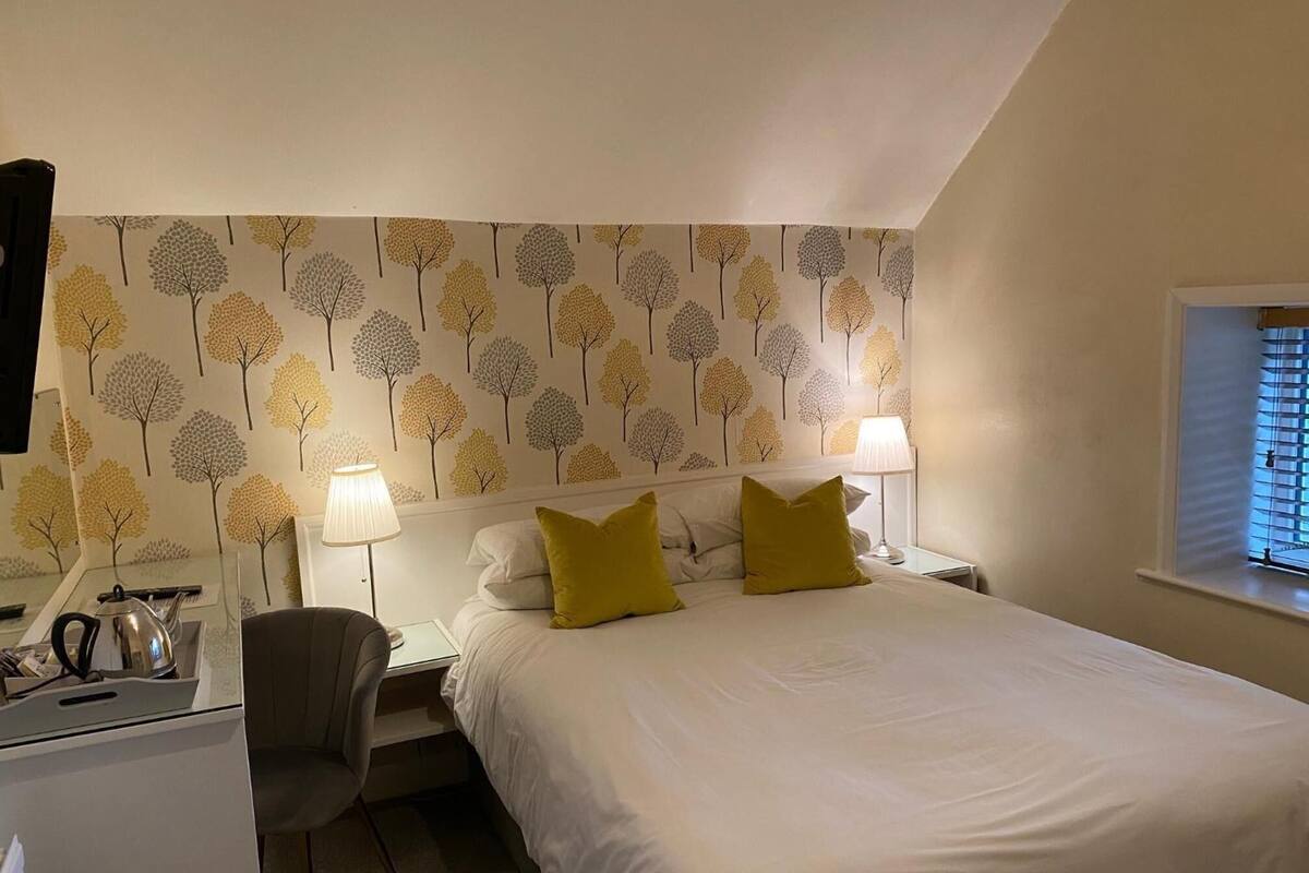 A bedroom with a bed, two bedside lamps, a desk with a kettle, and a chair.