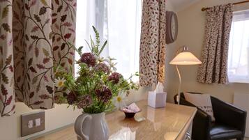 Una habitación con un escritorio de madera, un jarrón blanco con flores, una lámpara y una silla.