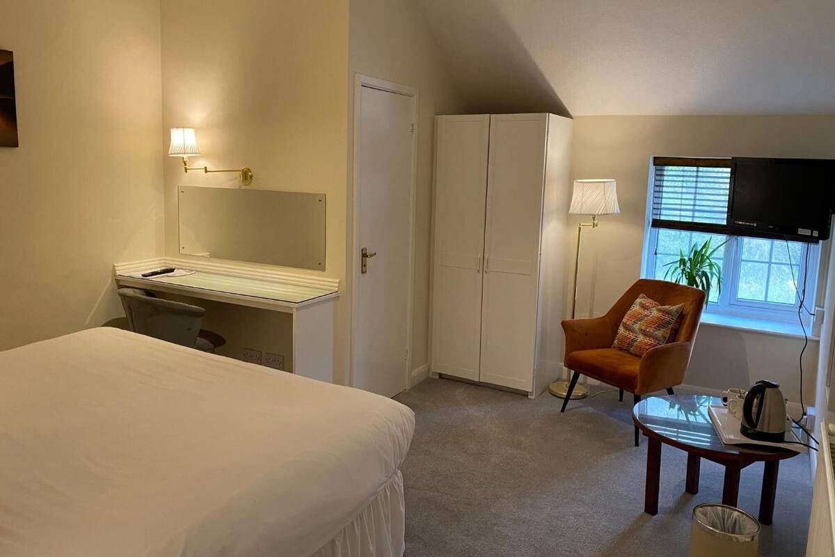 A hotel room with a bed, a desk with a mirror, a wardrobe, a chair, a small table with a kettle, and a TV mounted on the wall.