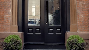 A black double door with a glass panel and a small window above it, set in a brick building with a small step leading up to the door. - The Broome (New York)