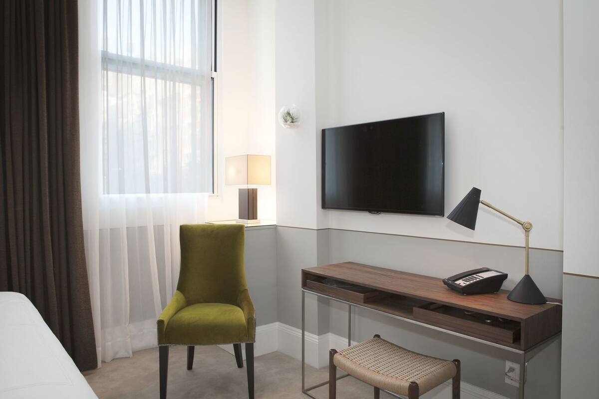 Hotellihuone, jossa on sänky, työpöytä lampun kanssa, tuoli, televisio ja ikkuna, jossa on läpikuultavat verhot.