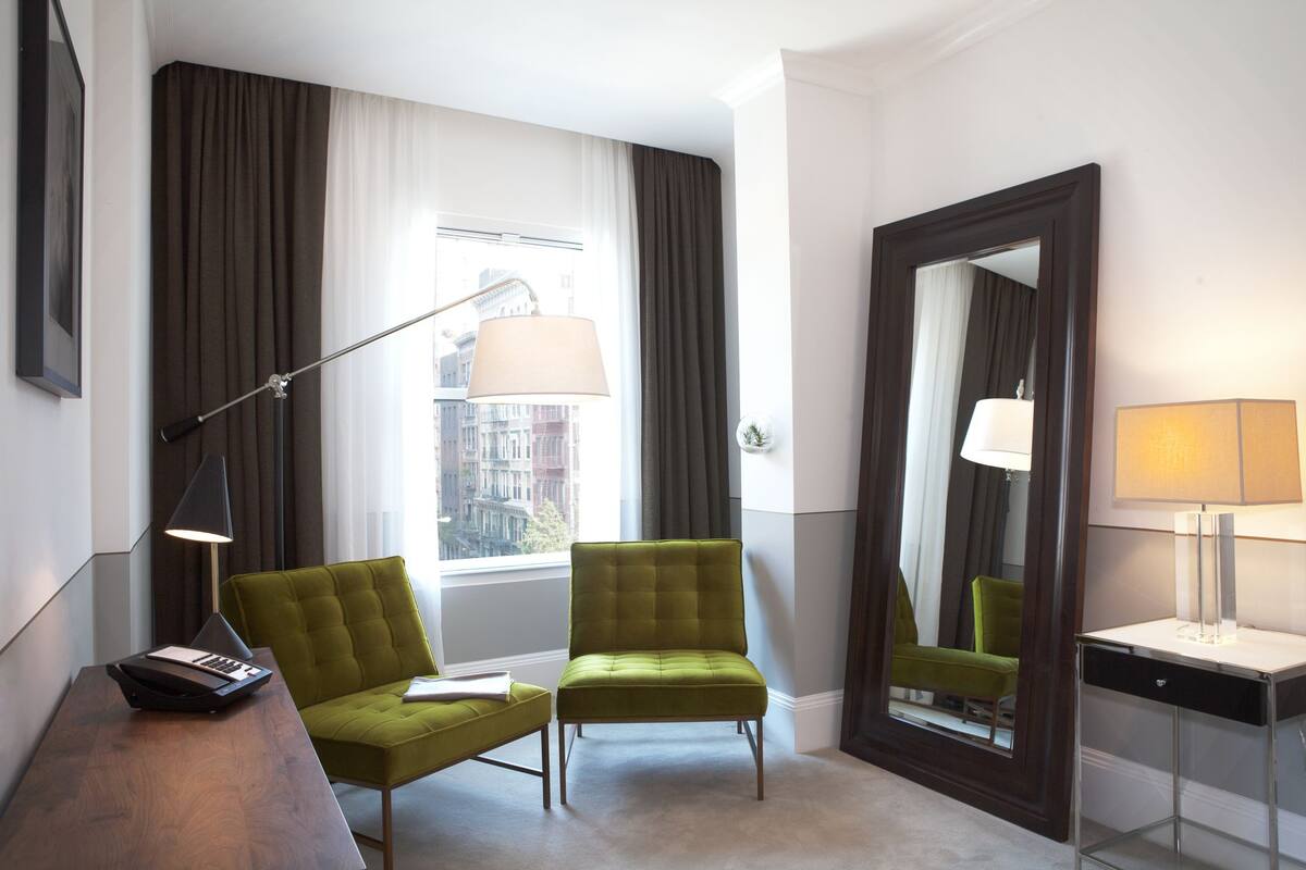Hotellihuone, jossa on vihreä nojatuoli, työpöytä lampun kanssa ja suuri peili.