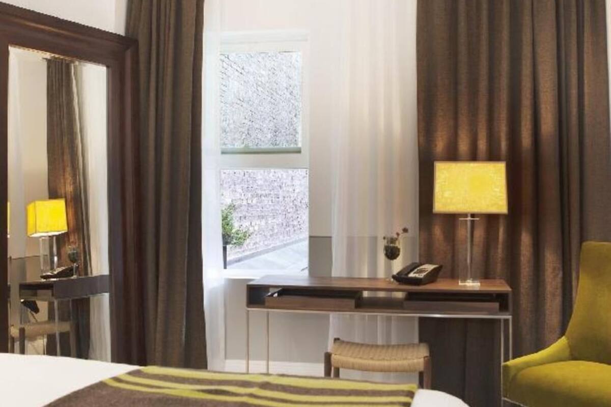 Hotellihuone, jossa on sänky, kirjoituspöytä lampun kanssa, tuoli ja ikkuna, jossa on verhot.