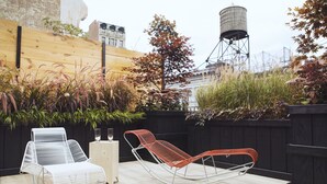 A rooftop patio with two lounge chairs, a small table, and a water tower in the background. - The Broome (New York)