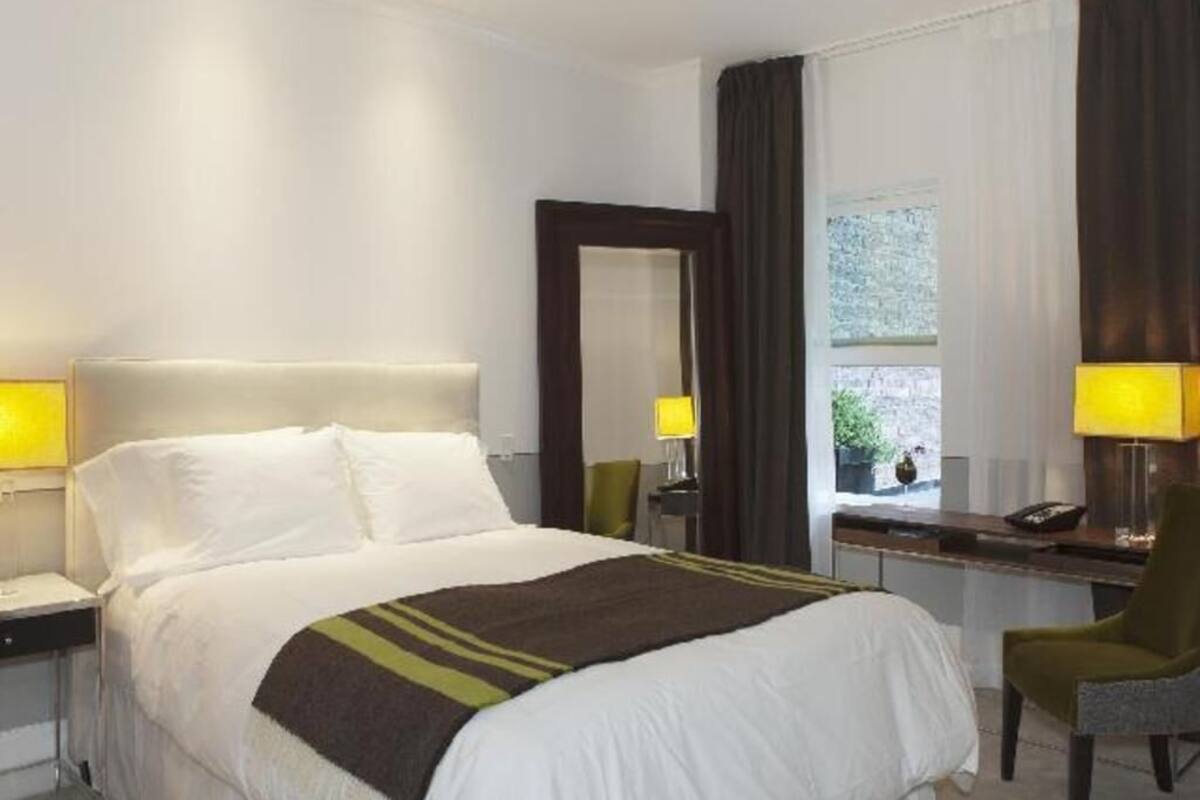 Hotellihuone, jossa on sänky, yöpöydät, työpöytä, tuoli, ikkuna verhoilla ja peili.