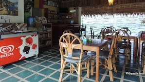 A restaurant with a tiled floor, wooden chairs, a table, a counter with a coffee machine, and a view of the sea through a window with a bamboo blind. - Asian Belgian Resort (Moalboal)