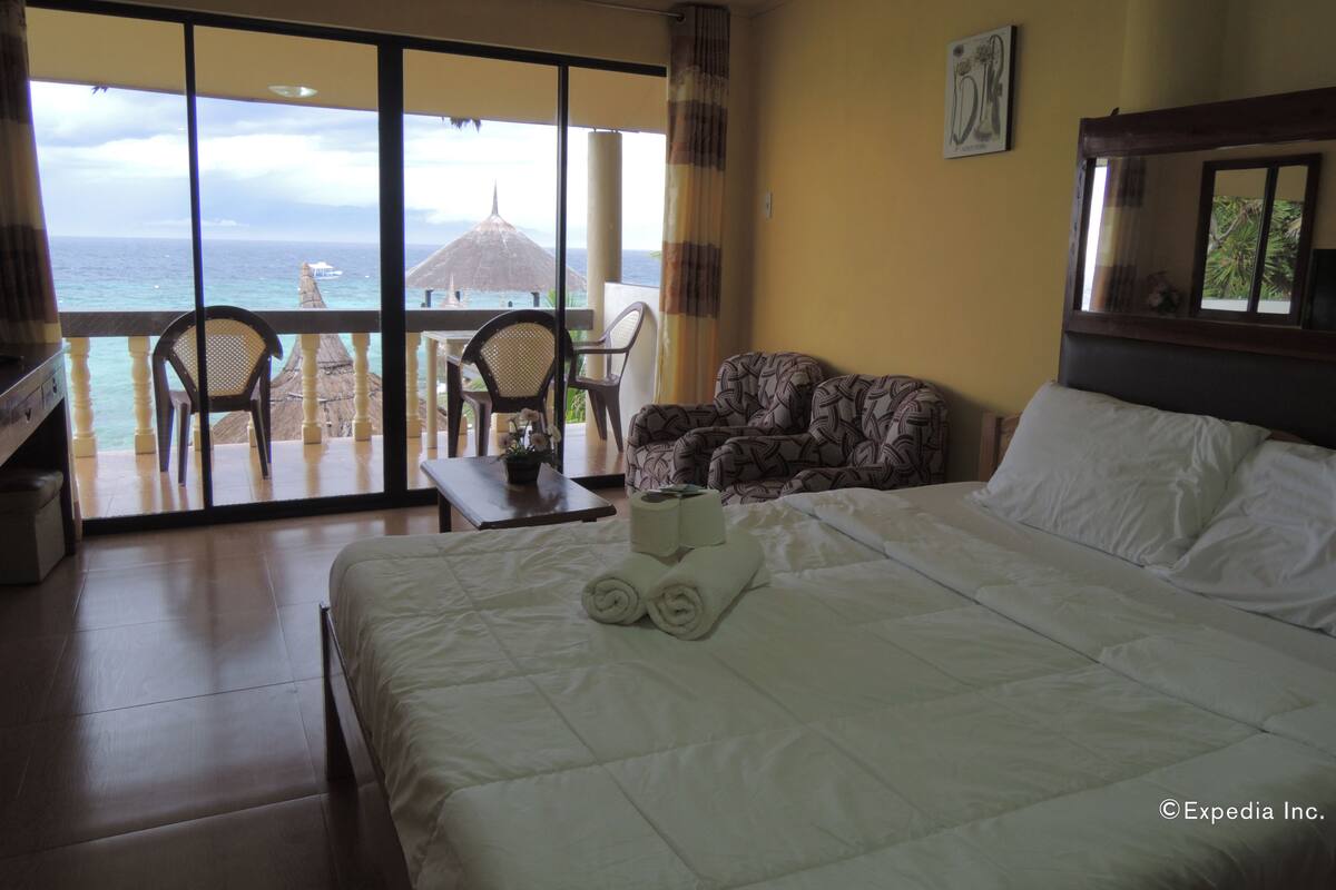 A hotel room with a bed, a small table, a chair, and a sofa. There is a large window with a view of the ocean and a thatched umbrella.