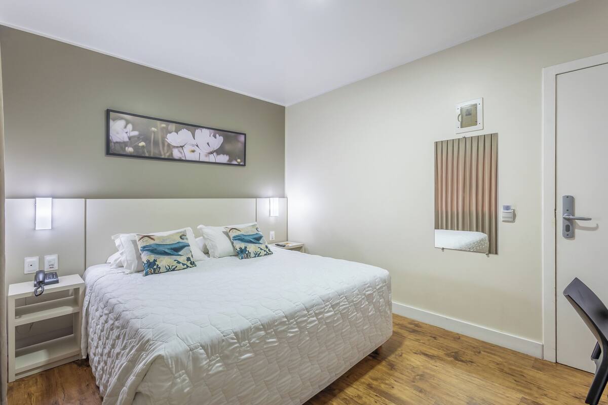 A neatly arranged bedroom with a bed, bedside tables, a nightstand, a wall-mounted light, a picture, a window with blinds, and a door.