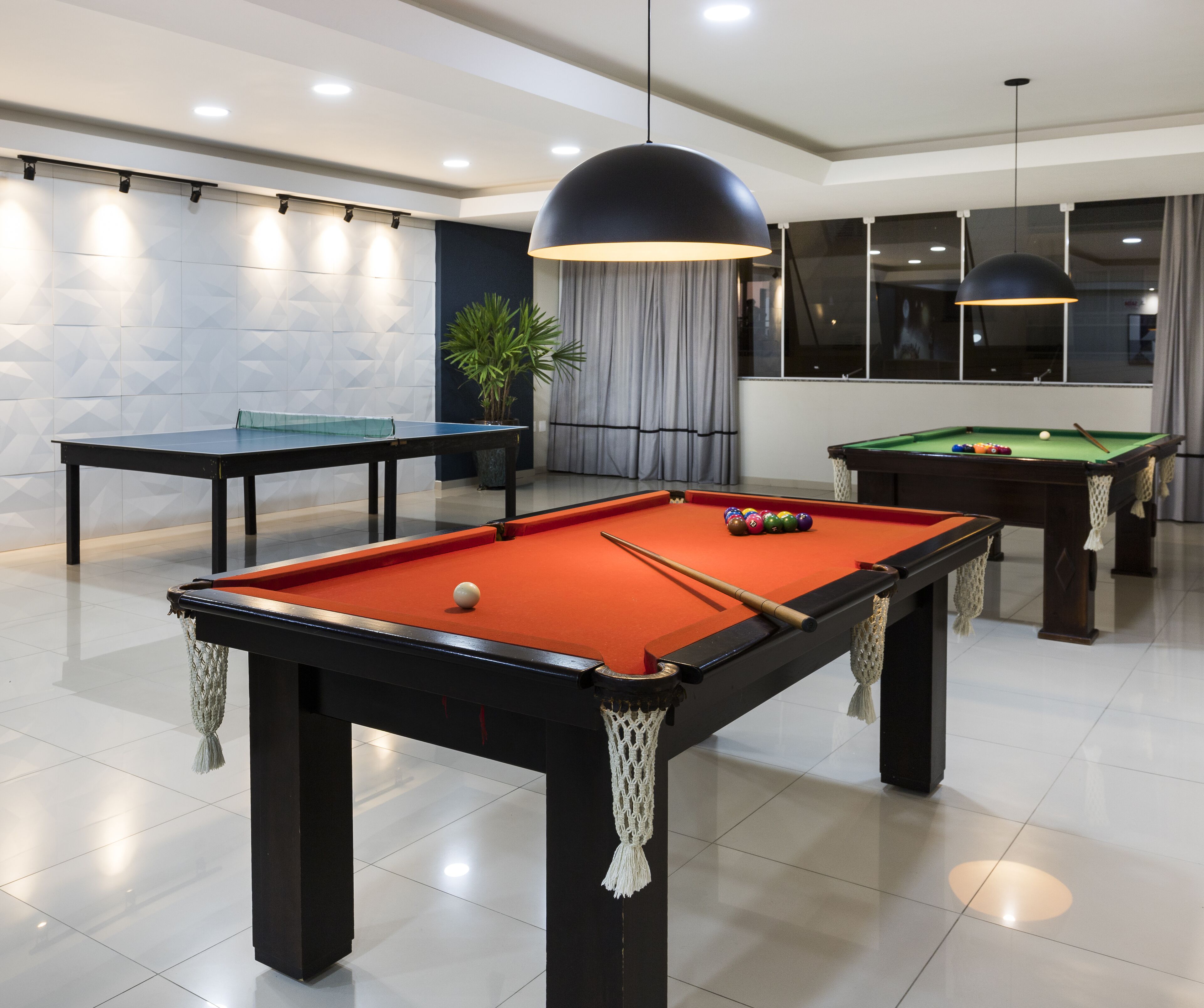 A pool table with balls, a billiards table, and a potted plant in a modern room with white walls and a large window.