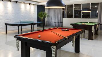 A pool table with balls, a billiards table, and a potted plant in a modern room with white walls and a large window.