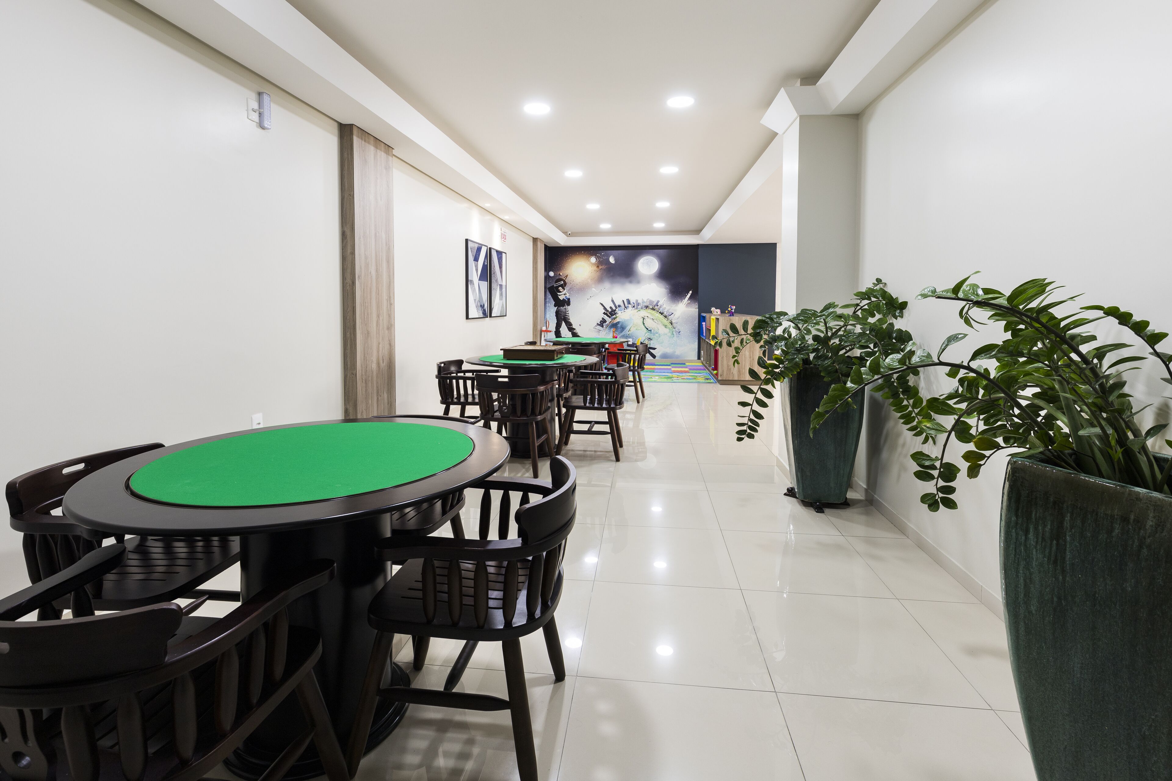 Una habitación con una mesa de póker verde, sillas negras, una planta grande en maceta y un mural en la pared.