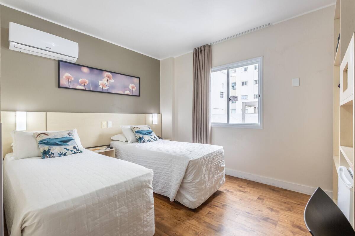 A hotel room with two beds, a window with curtains, a painting on the wall, and an air conditioning unit.