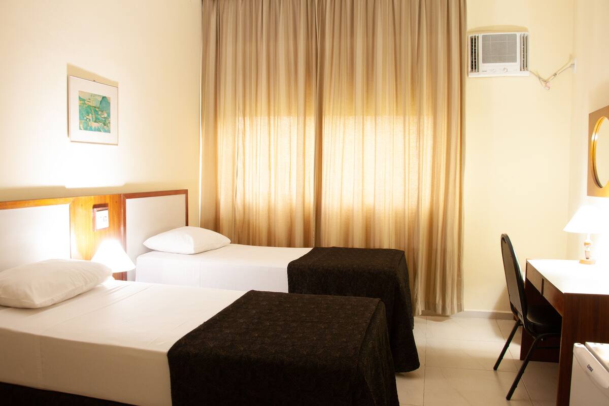 Habitación de hotel con dos camas, un escritorio con silla, lámpara, equipo de aire acondicionado y ventana con cortinas.