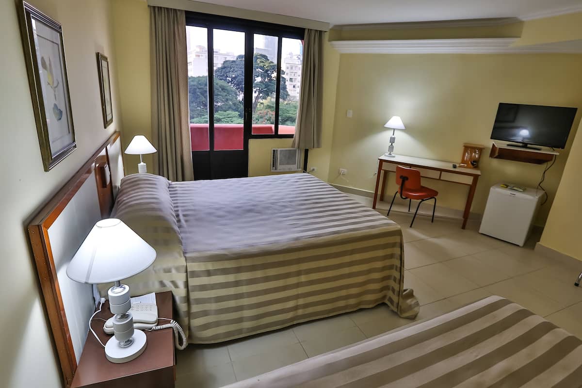 Habitación de hotel con cama, mesita de noche con lámpara, escritorio con silla, televisor y ventana con vista a edificios y árboles.