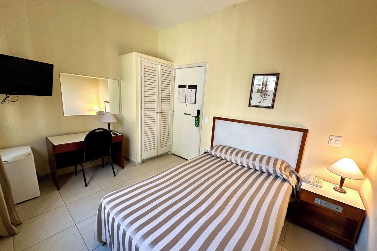 Una habitación de hotel con cama, escritorio, silla, televisión, armario y mesita de noche con lámpara.