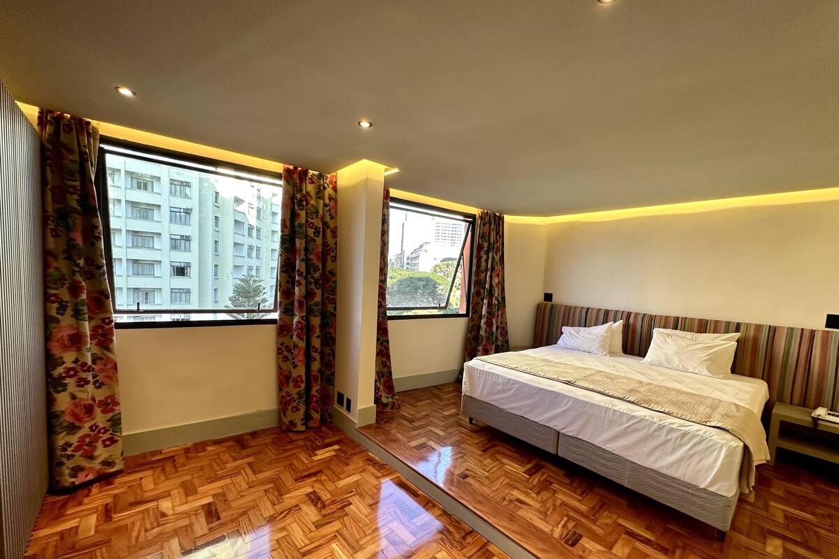 Habitación de hotel con una cama grande, suelo de parqué de madera, dos ventanas con cortinas estampadas y vistas a un edificio.