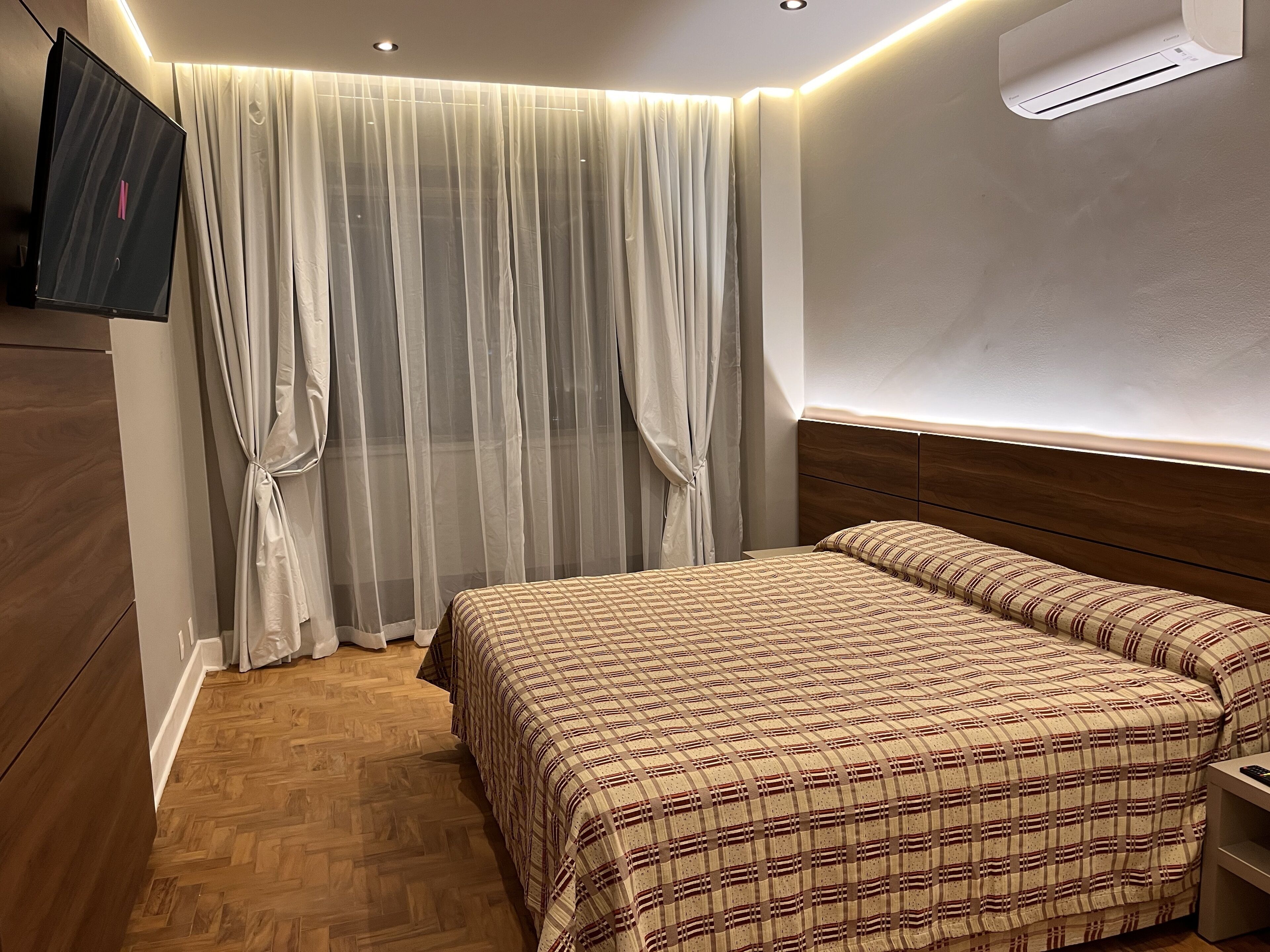 A bedroom with a bed, curtains, an air conditioning unit, and a television.