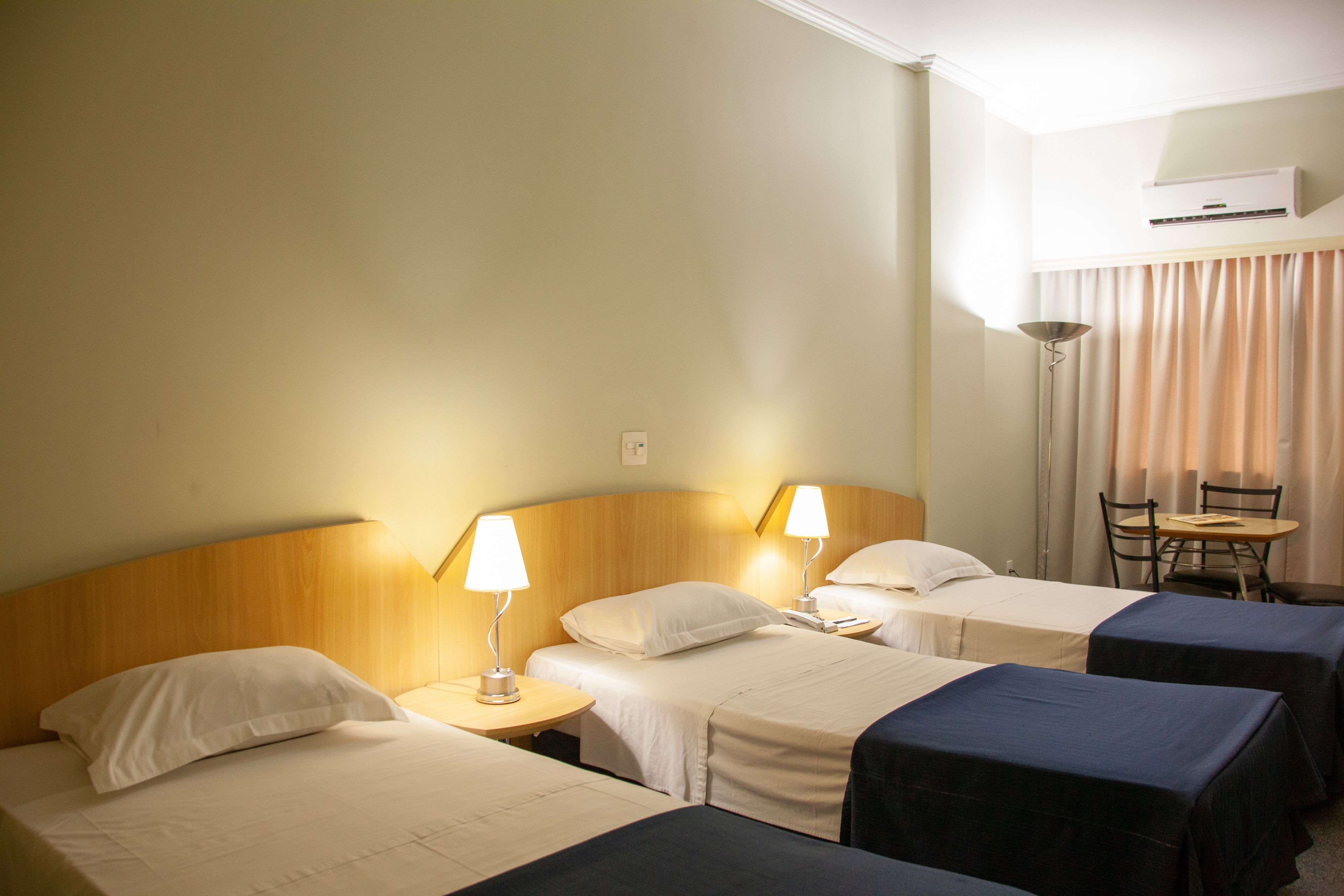 A hotel room with three beds, each with a bedside table and lamp, a small table with chairs, and an air conditioning unit on the wall.