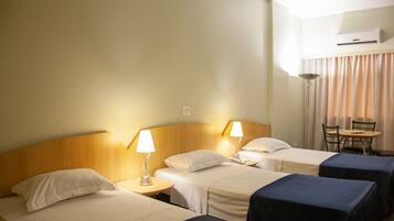 A hotel room with three beds, each with a bedside table and lamp, a small table with chairs, and an air conditioning unit on the wall.