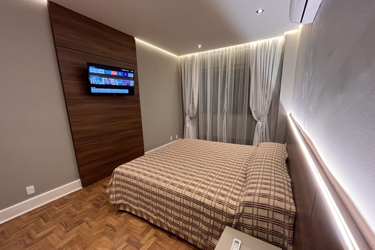 Une chambre avec un plancher en bois, un lit recouvert d’une couverture à carreaux, un téléviseur fixé sur un panneau en bois et une table de chevet avec une télécommande.