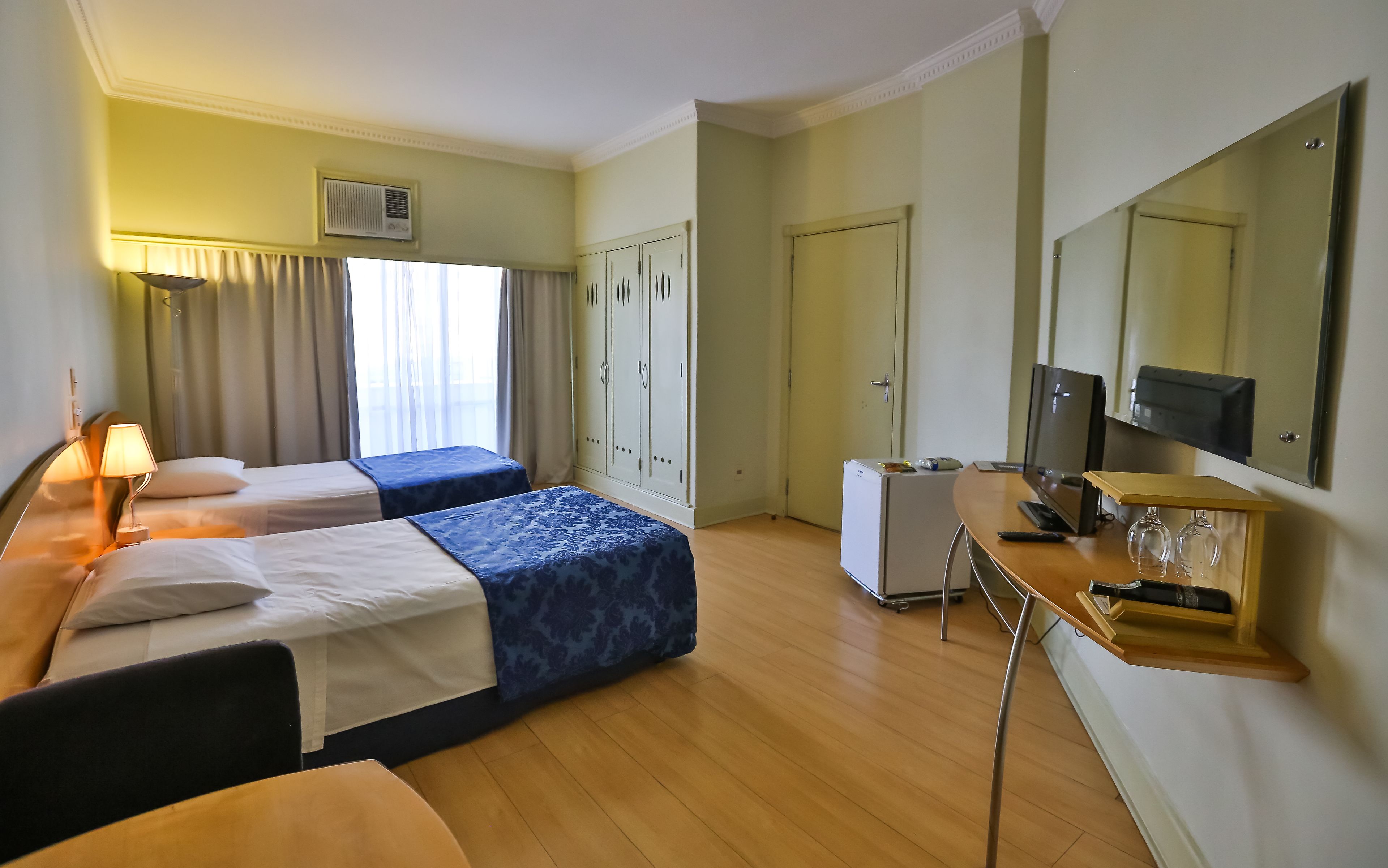 A hotel room with two beds, a desk, a mirror, a television, and a minibar.