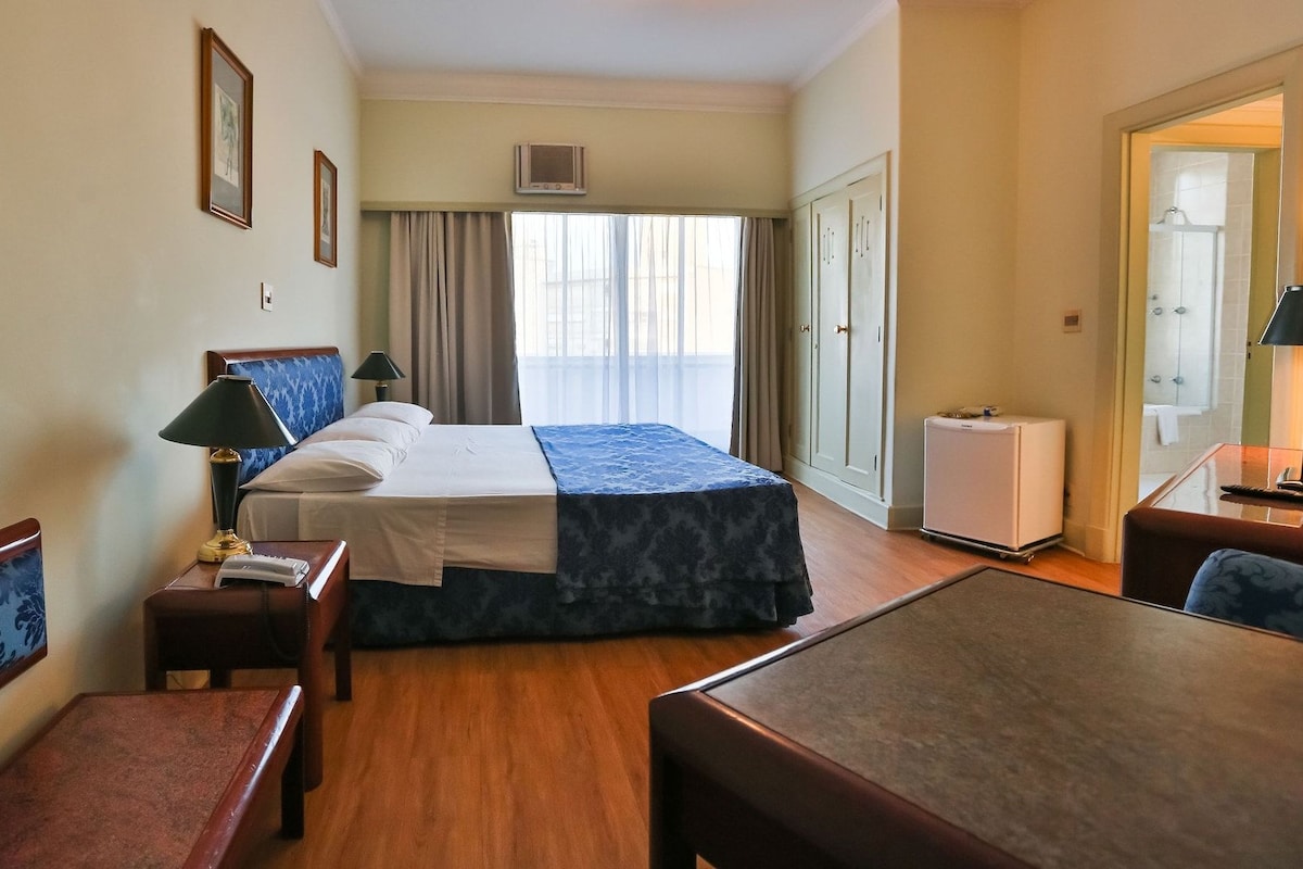 Habitación de hotel con cama, mesitas de noche, escritorio, silla, armario y baño visible a través de una puerta abierta.