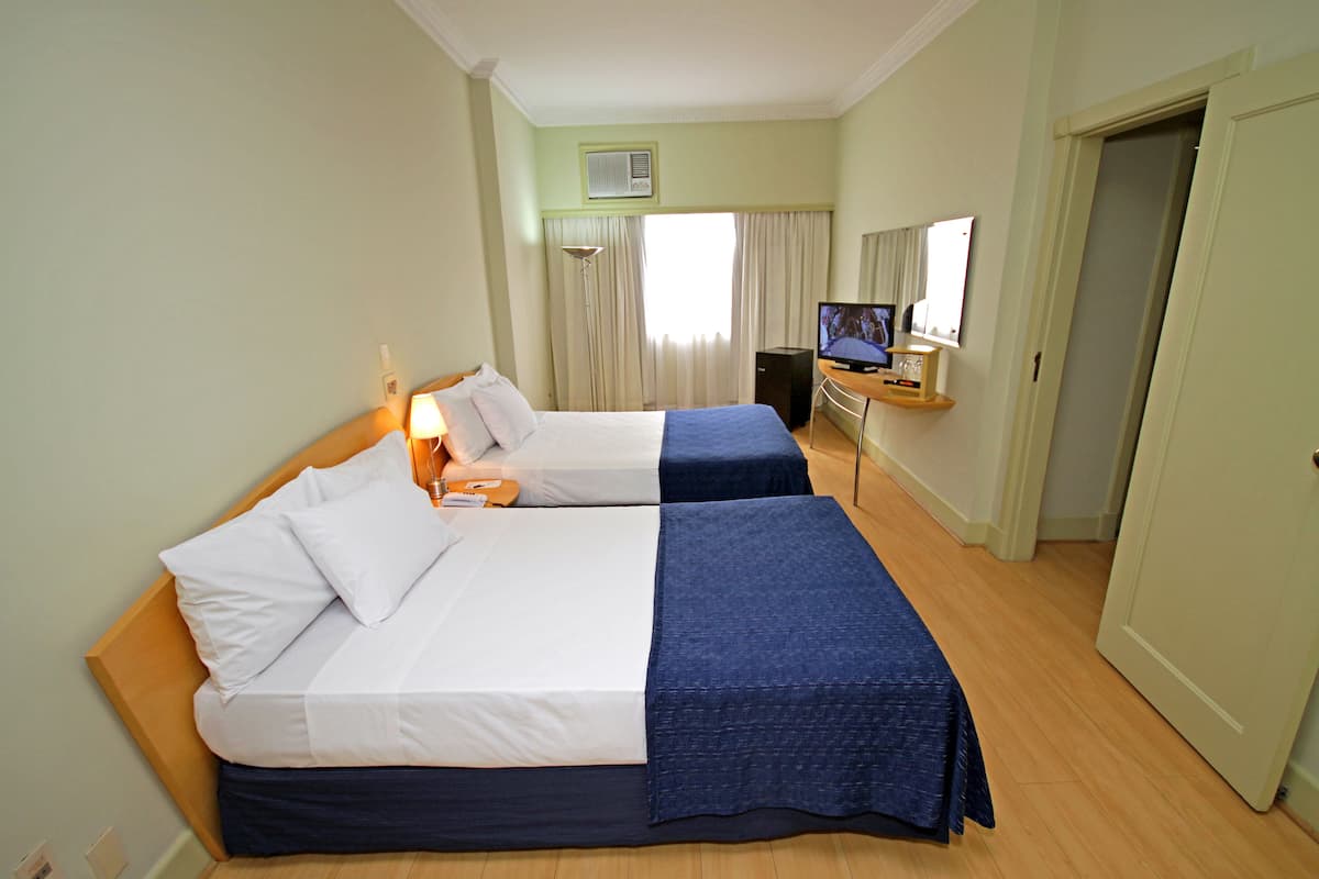Une chambre d’hôtel avec deux lits, un bureau avec un ordinateur, une télévision et une fenêtre avec des rideaux.