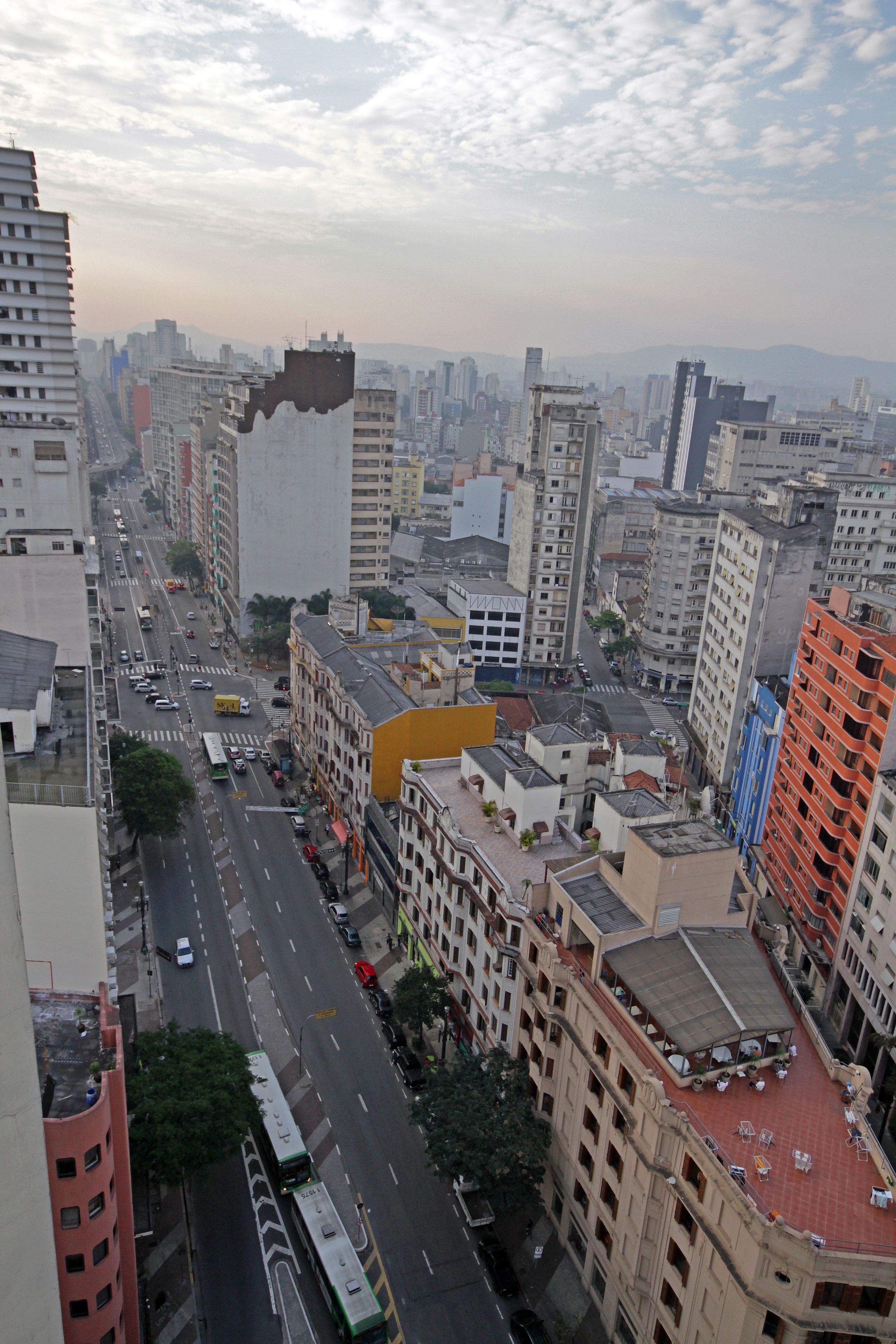 Paisagem urbana com edifícios altos, rua movimentada e área no terraço com mesas ao ar livre.