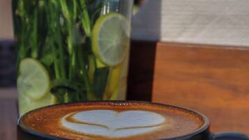 Una jarra de vidrio transparente con hierbas verdes y rodajas de lima, una taza negra con arte de latte, y una caja de madera sobre una mesa.