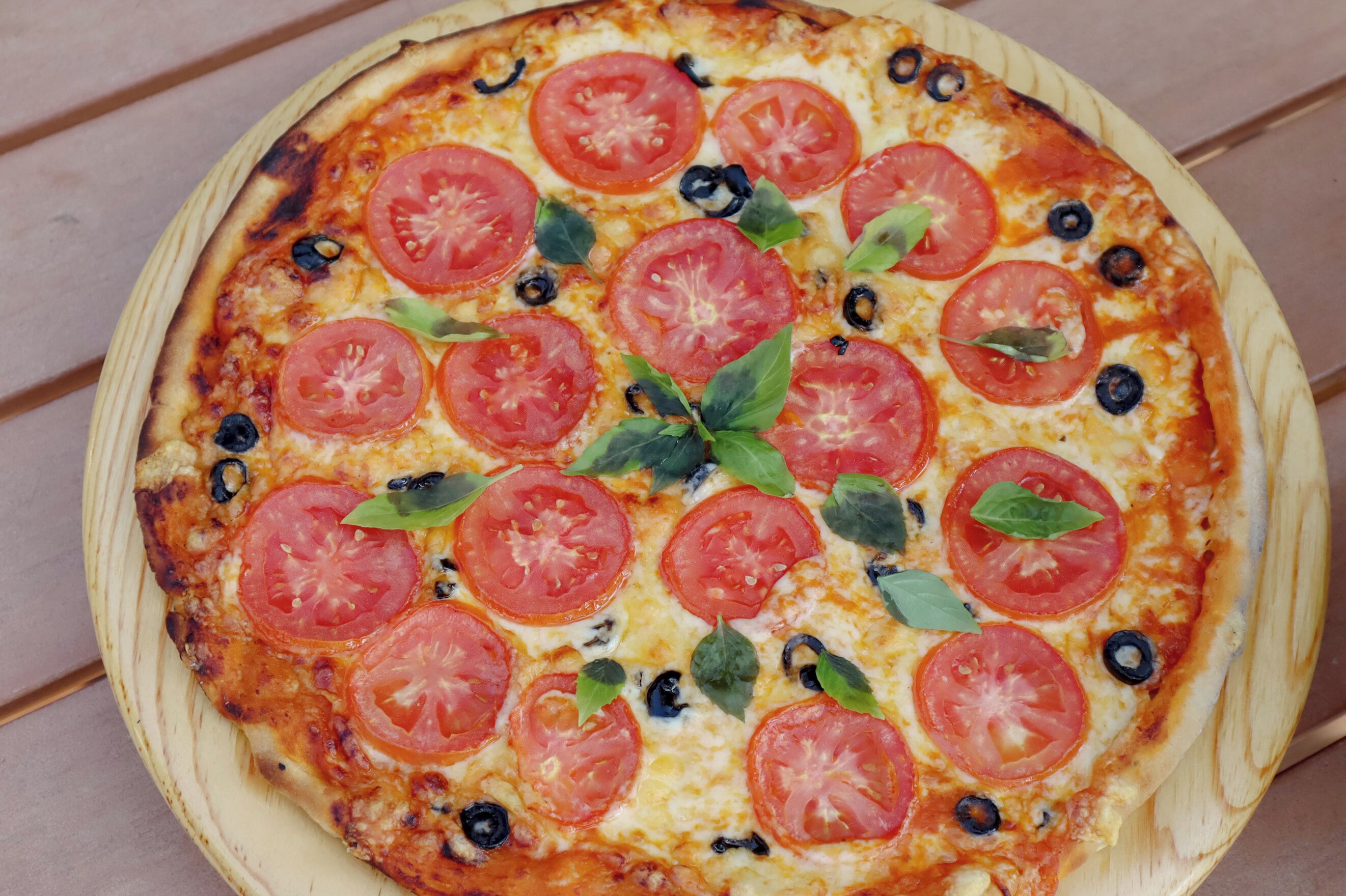 Pizza con rodajas de tomate, aceitunas y hojas de albahaca sobre una tabla de madera.