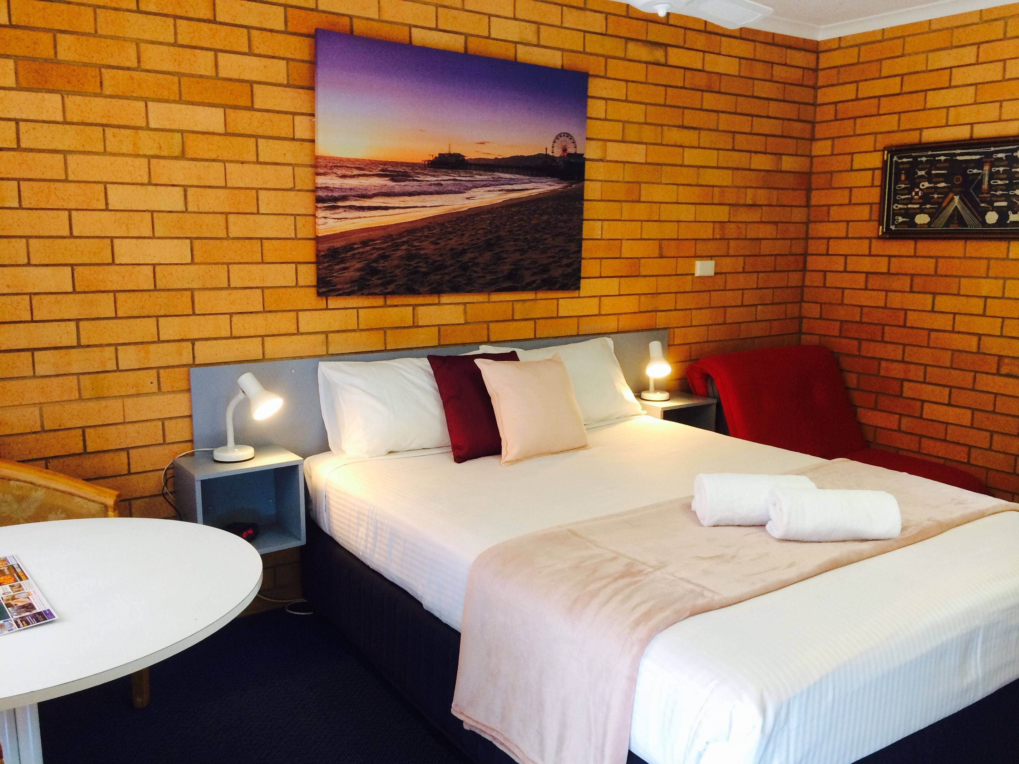 A hotel room with a brick wall, a bed with white and red bedding, a nightstand with a lamp, a red chair, and a round table with a magazine.