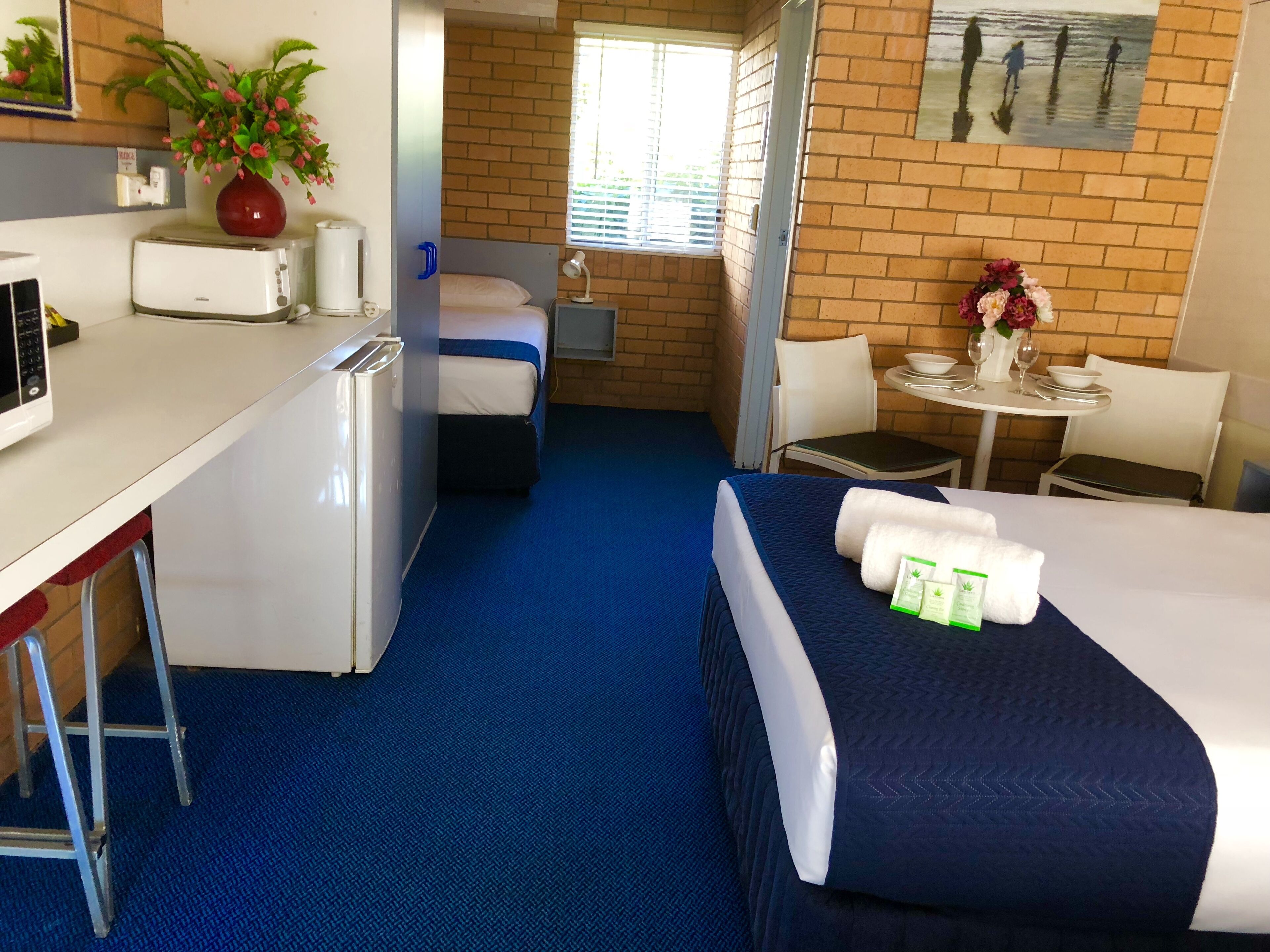 Habitación de hotel con cama, escritorio con microondas, pequeña mesa con un florero y una silla, además de ventana con vista a personas a lo lejos.