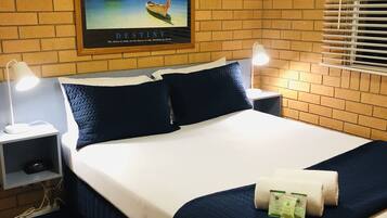 Une chambre d’hôtel avec un mur en briques, un lit avec une literie blanche et bleu marine, deux tables de chevet avec des lampes, un tableau encadré au mur et une fenêtre avec des stores.