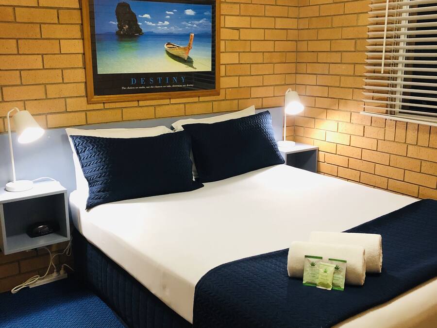 Une chambre d’hôtel avec un mur en briques, un lit avec une literie blanche et bleu marine, deux tables de chevet avec des lampes, un tableau encadré au mur et une fenêtre avec des stores.