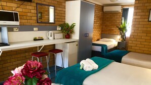 Une chambre d’hôtel avec une kitchenette, un lit recouvert d’une courtepointe turquoise, un petit bureau avec une chaise, un miroir et une plante en pot.