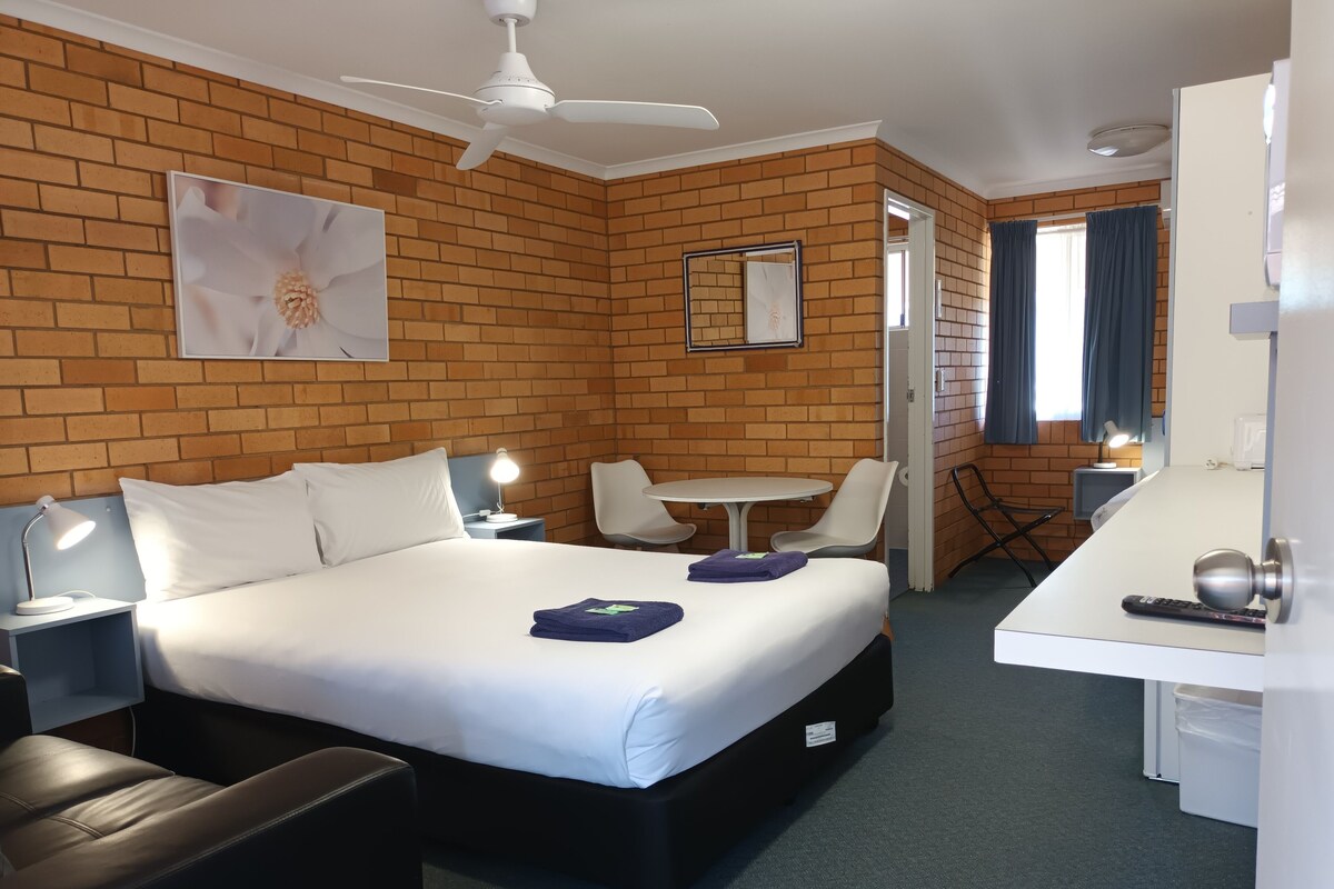 Habitación de hotel con cama, escritorio, silla, mesa, ventilador de techo y pared de ladrillo.