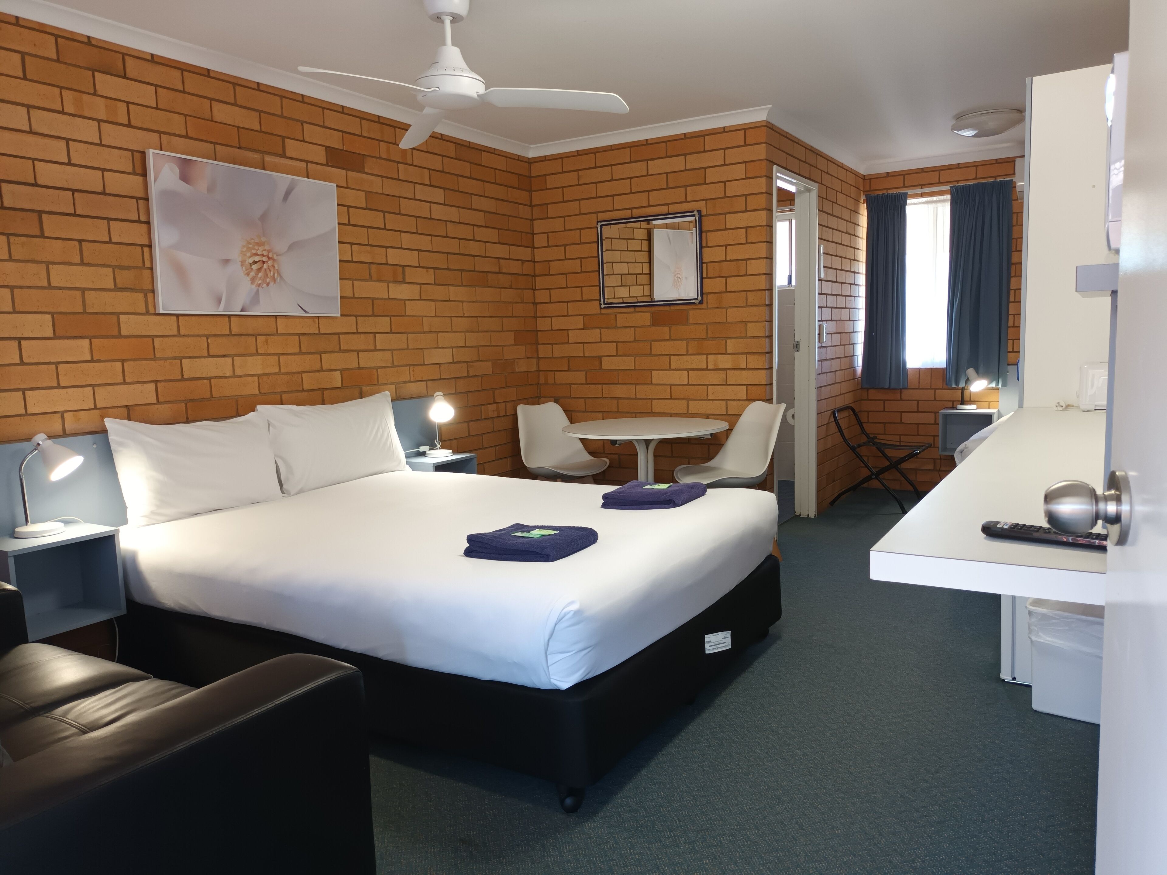 A hotel room with a brick wall, a bed with white linens, a desk with a chair, a small table, and a ceiling fan.