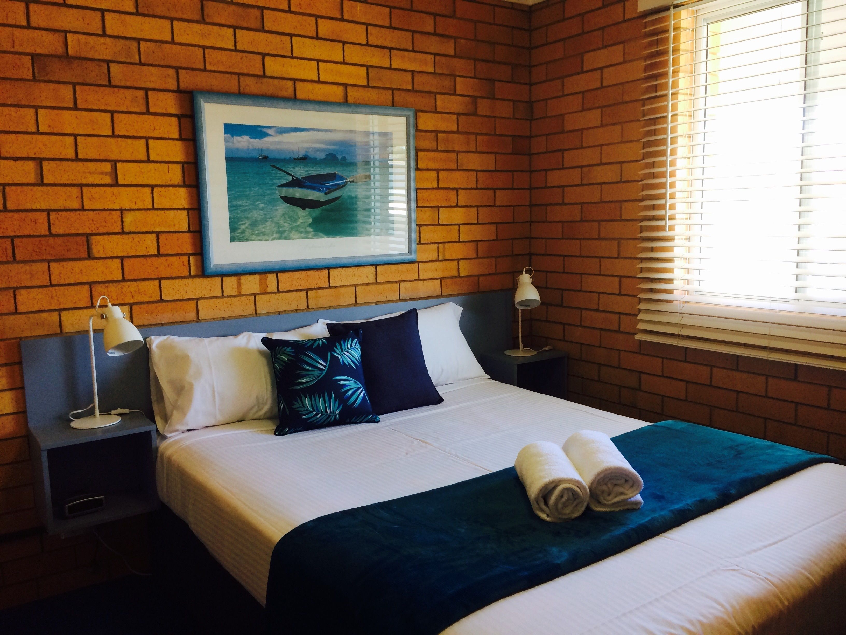 A hotel room with a brick wall, a framed picture of a boat, a neatly made bed with pillows, and two bedside lamps.