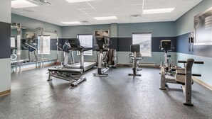Academia com diversos equipamentos de exercício, janelas amplas e um espelho grande na parede.