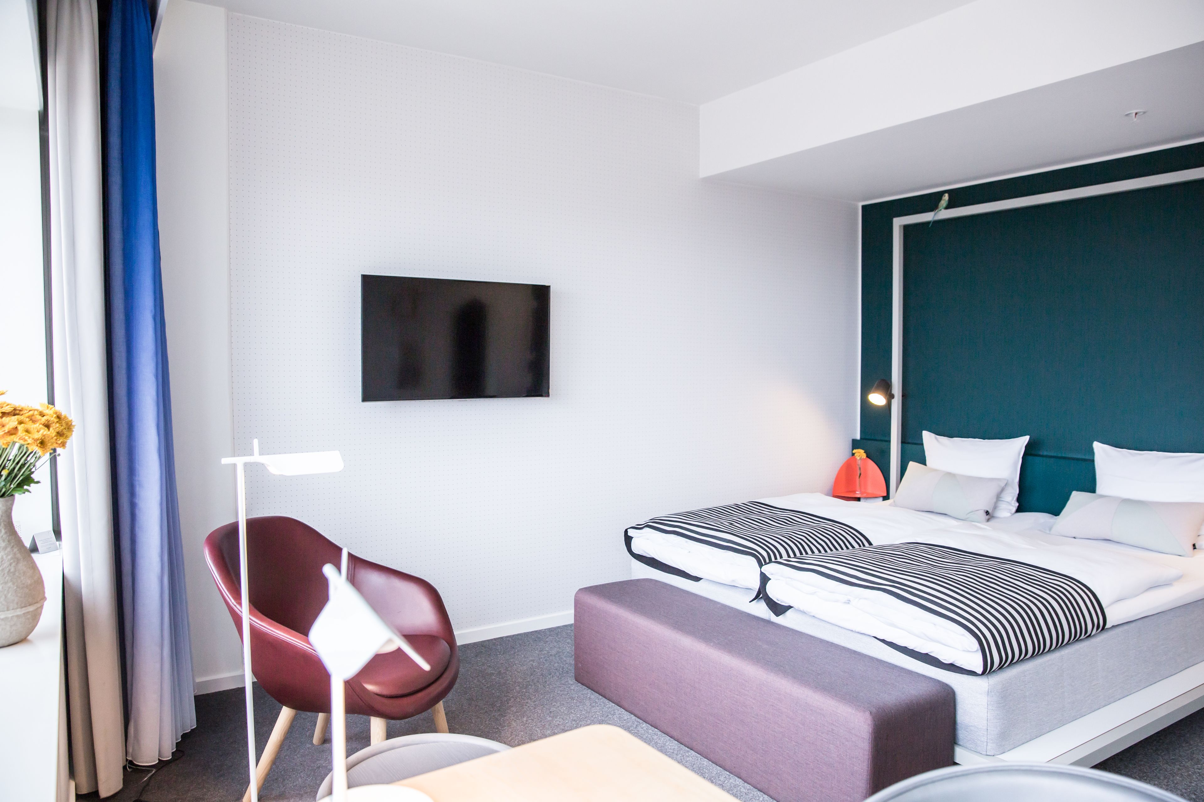 A modern hotel room with a large bed, a TV mounted on the wall, a red chair, and a small table.